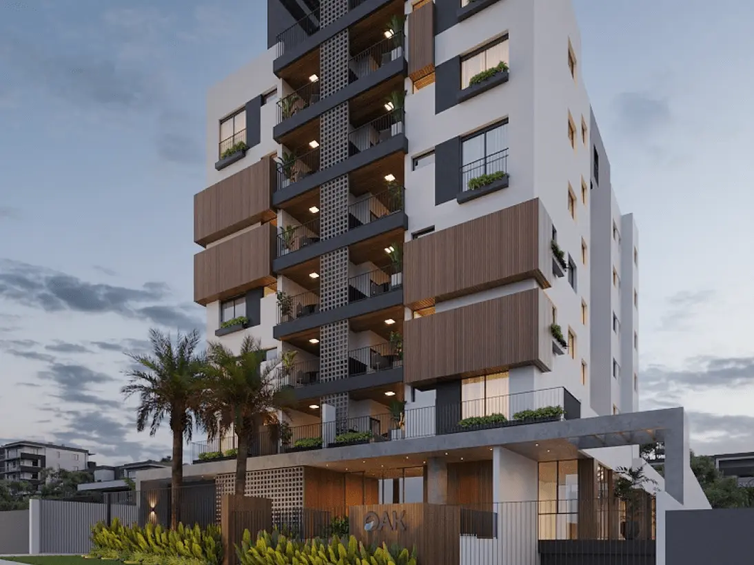 Um edifício residencial alto e moderno, chamado "Oak", com design assimétrico, varandas com detalhes em madeira e iluminação suave ao entardecer. A entrada apresenta o nome "OAK" em destaque e é cercada por vegetação e palmeiras.
